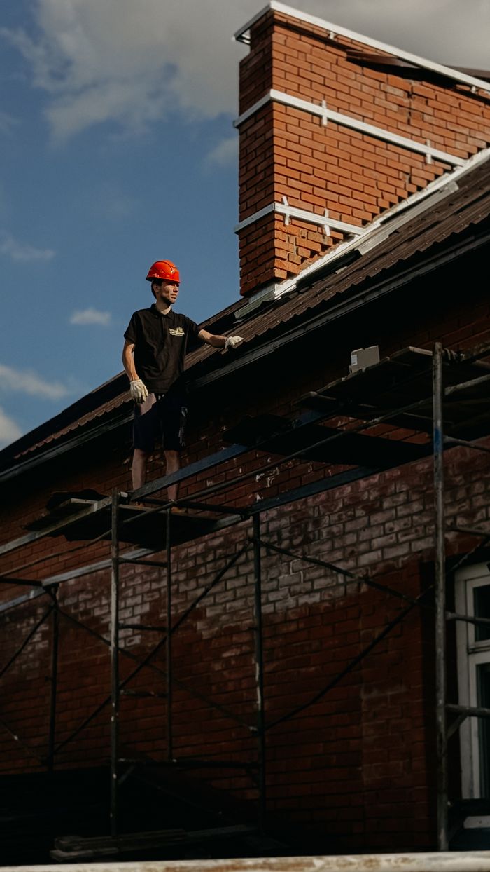 Construction worker stands on scaffolding next to a brick chimney, wearing safety gear.